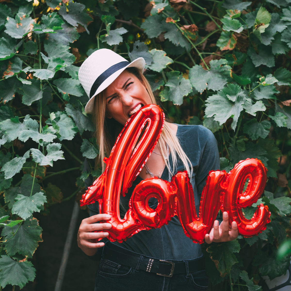 Une femme coiffée d'un chapeau mord dans un ballon gonflable rouge en forme du mot « love », devant un feuillage vert.