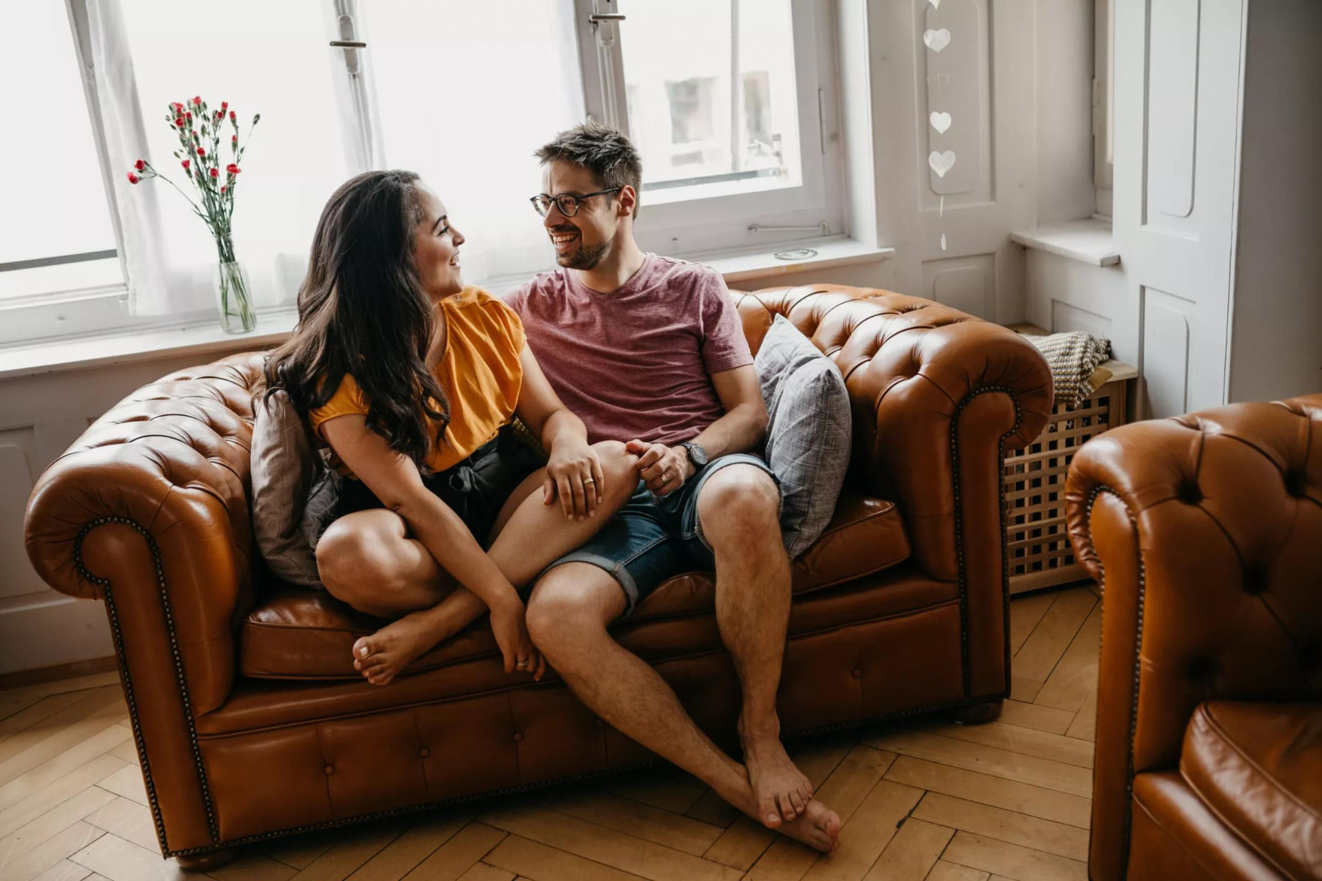 Un couple assis sur un canapé en cuir dans un salon lumineux, en train de rire et de se regarder.